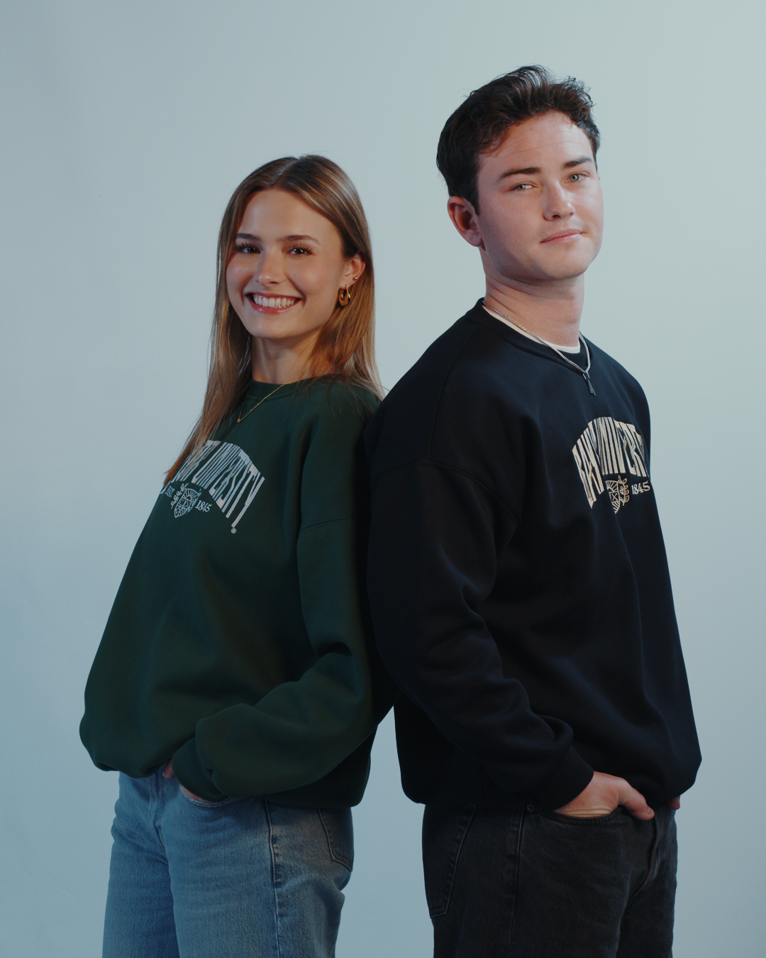 Two models wearing crewneck sweaters, one in dark green and the other in black, with embroidered text on the front. Baylor University