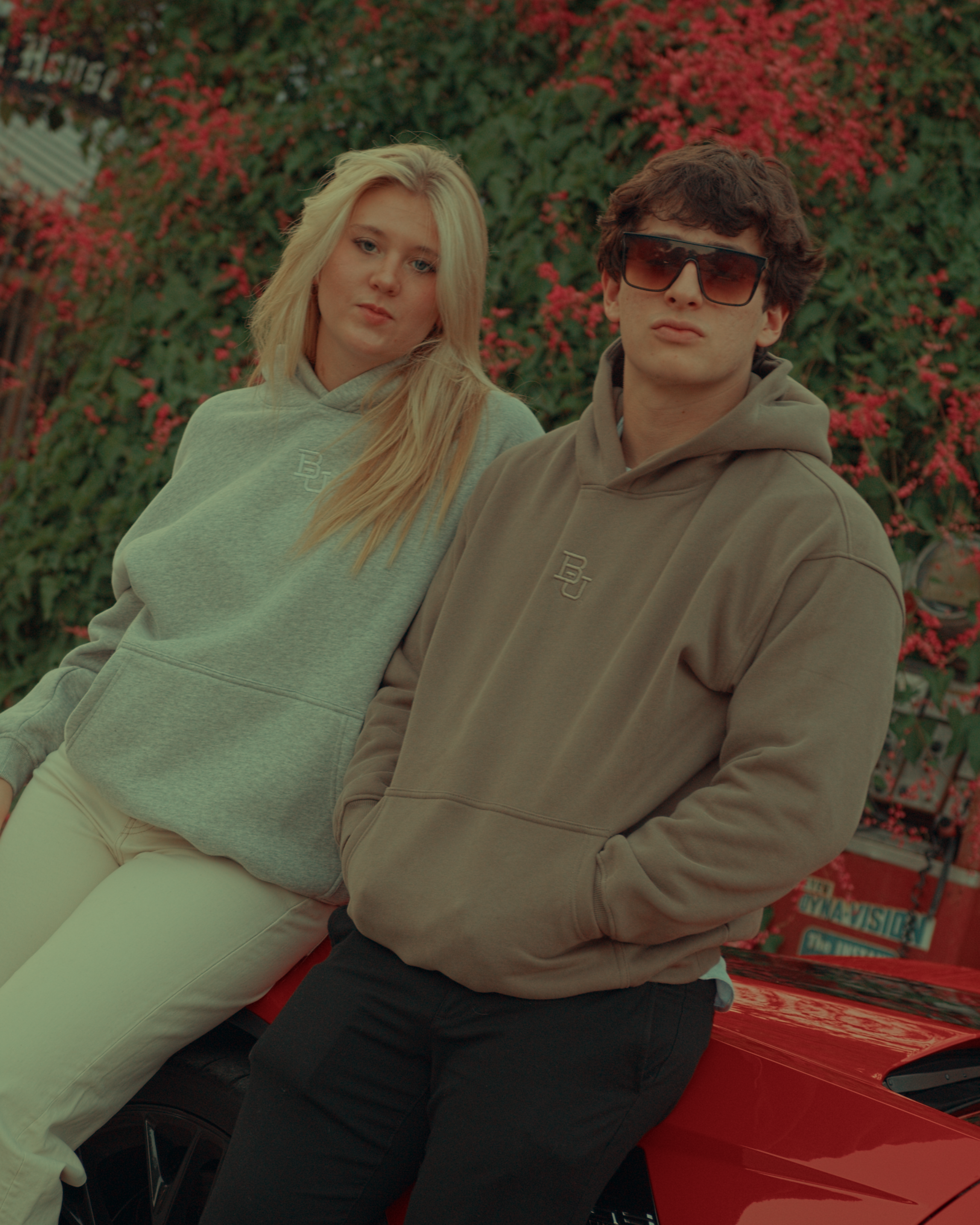 Two people sitting on a red car with green leaves background wearing Baylor University hoodies