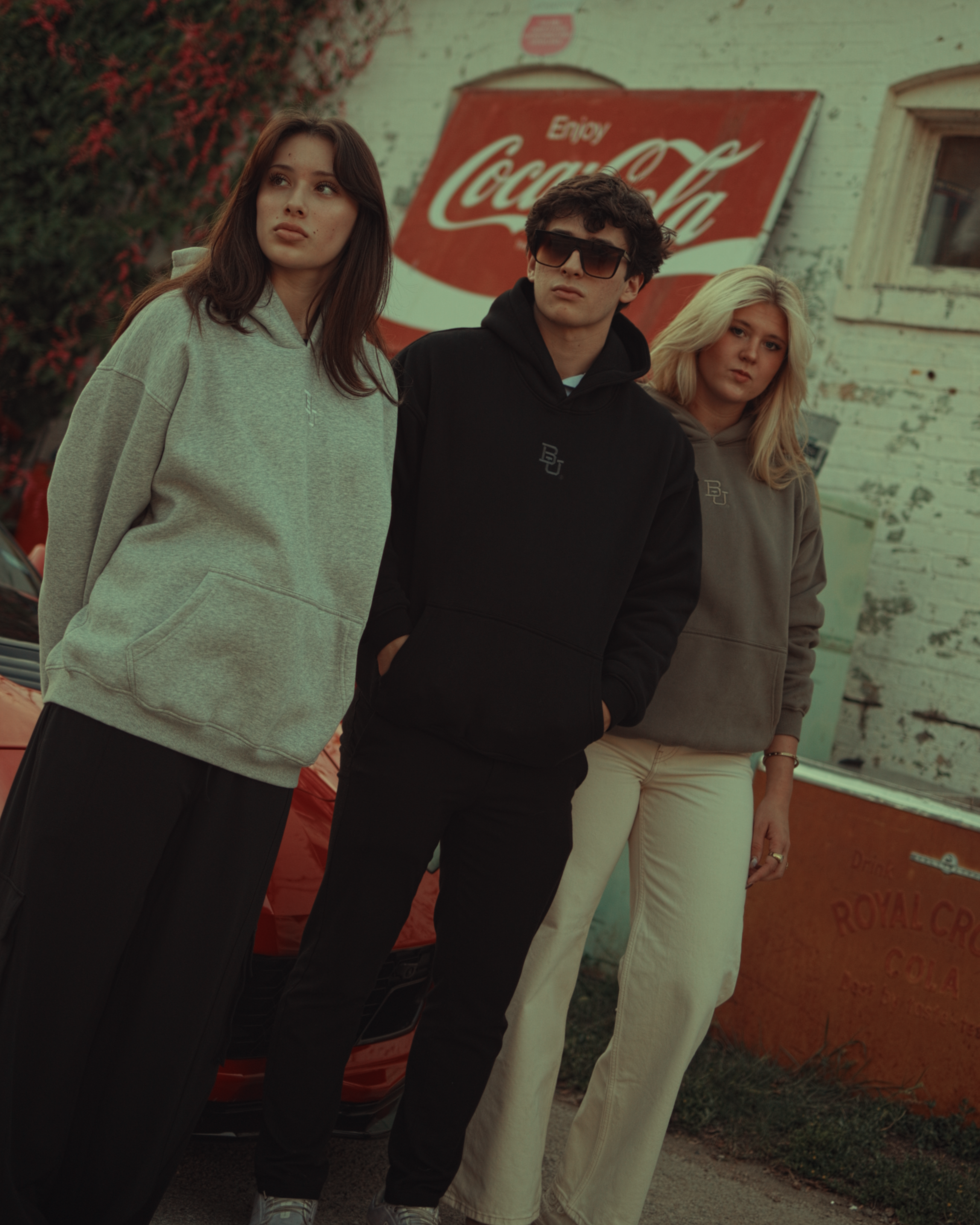 Three people standing in front of a Coca-Cola sign. Wearing Mclane Stadium Hoodies