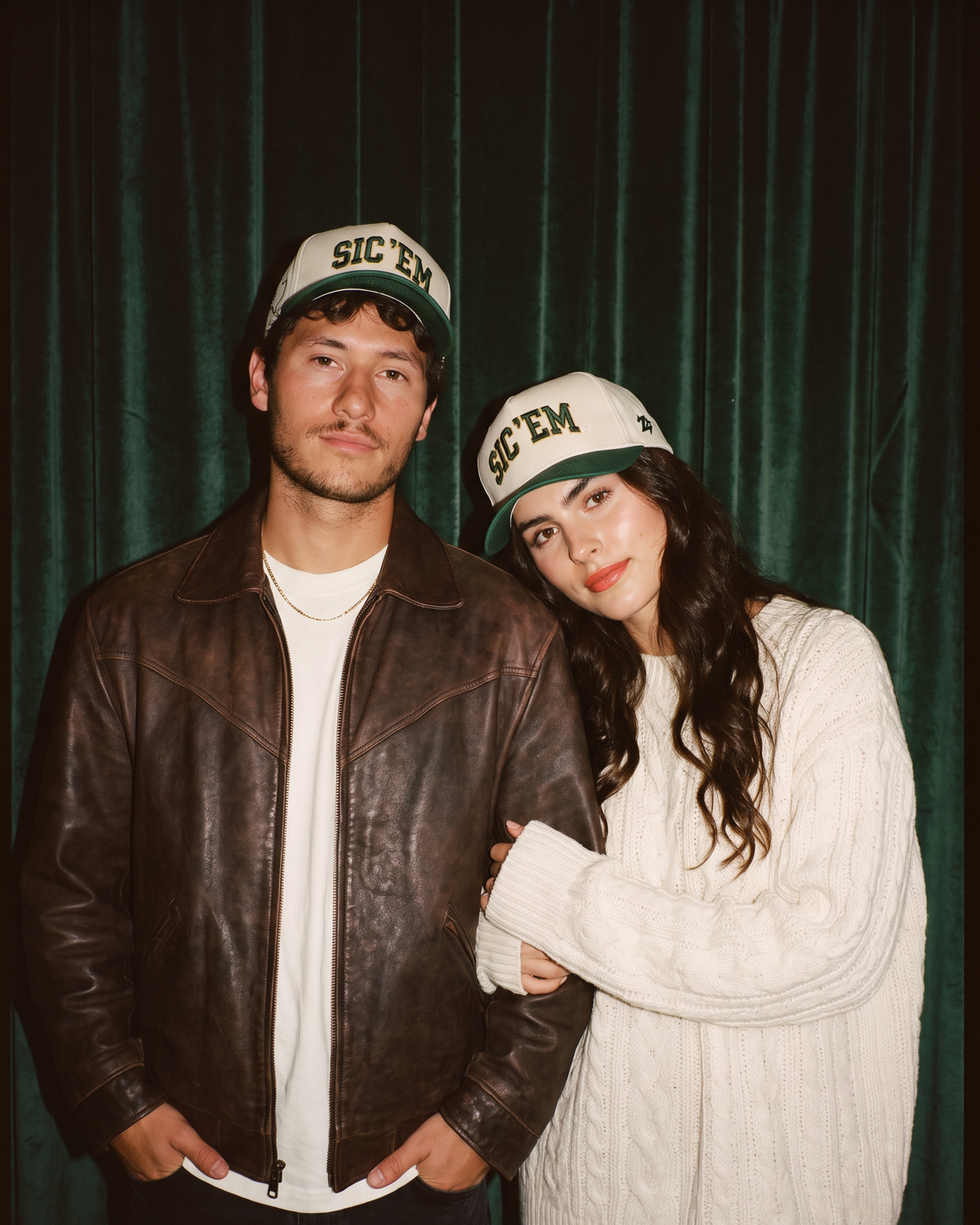 A man and a woman posing together in front of a dark green velvet curtain. Both are wearing matching beige and green 'SIC 'EM' baseball caps, with the man wearing a brown leather jacket over a white t-shirt and the woman wearing a cream cable-knit sweater.