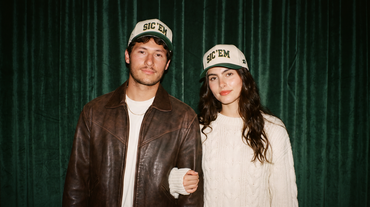A man and a woman standing in front of a dark green velvet curtain, both wearing matching white and green 'SIC 'EM' baseball caps. The man wears a brown leather jacket over a white shirt, and the woman wears a cream cable-knit sweater.