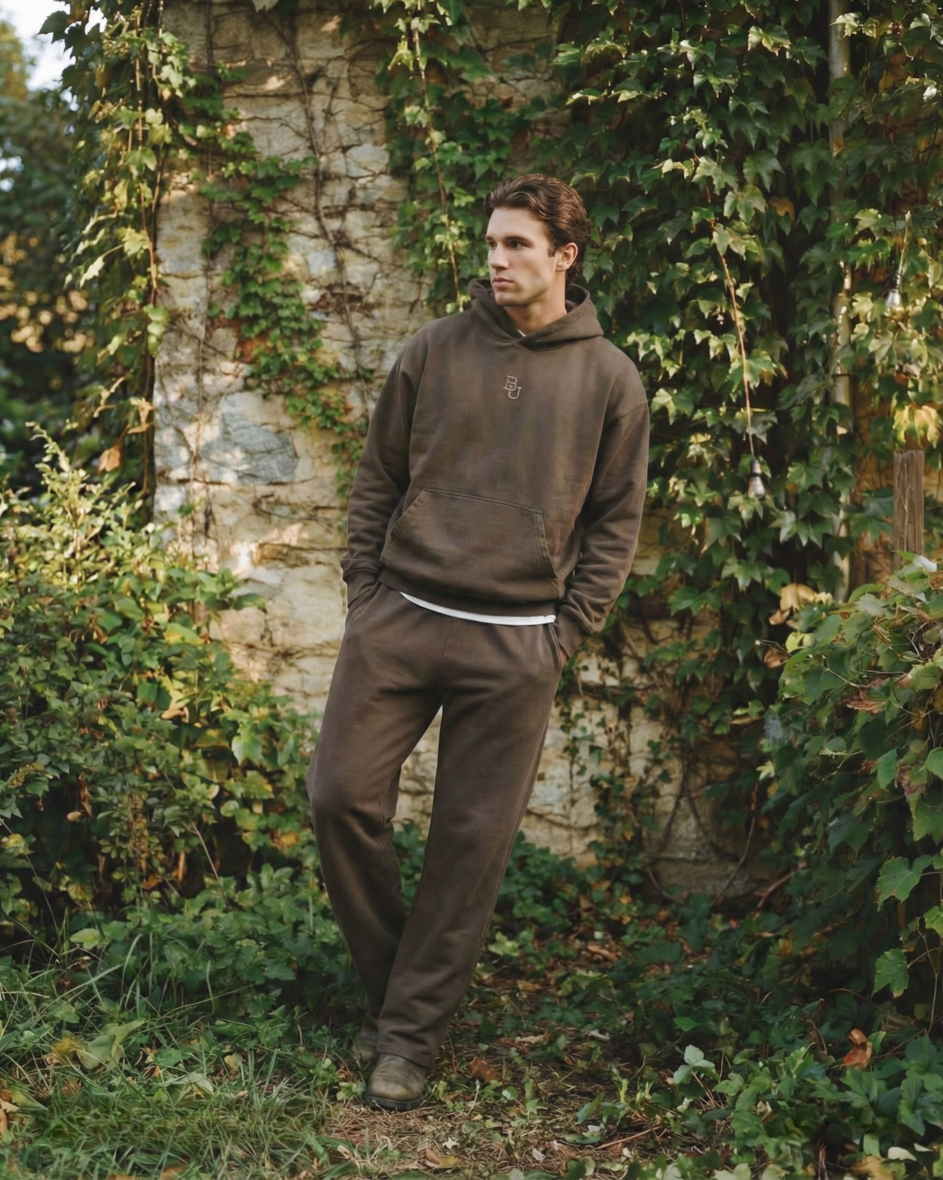 Man wearing a brown baylor university hoodie and pants standing in front of a stone wall with greenery.