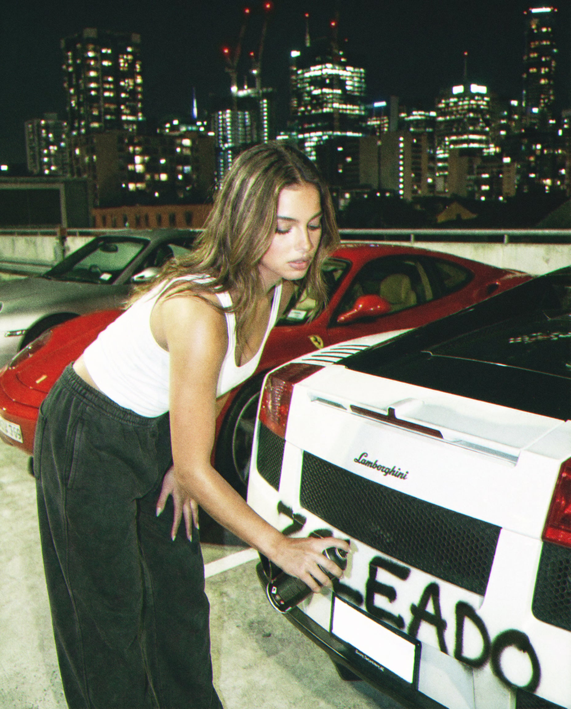 Woman standing next to a white Lamborghini with a city skyline in the background, she spray paitning "zoleado"