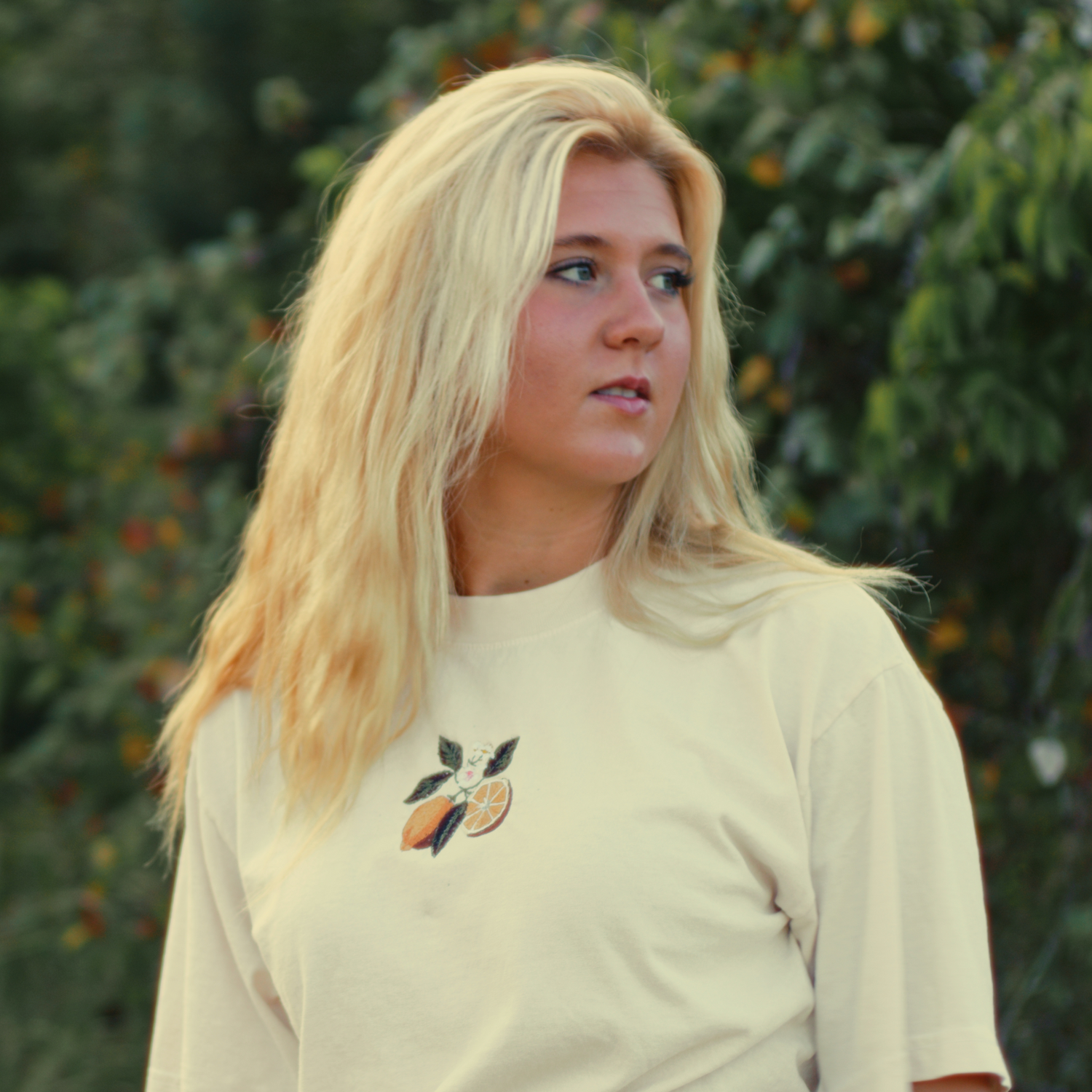 Woman wearing an embroidered lemon-themed shirt, standing outdoors in a natural setting. Blonde girl wearing a lemon shirt. Girl wearing a white shirt in nature.