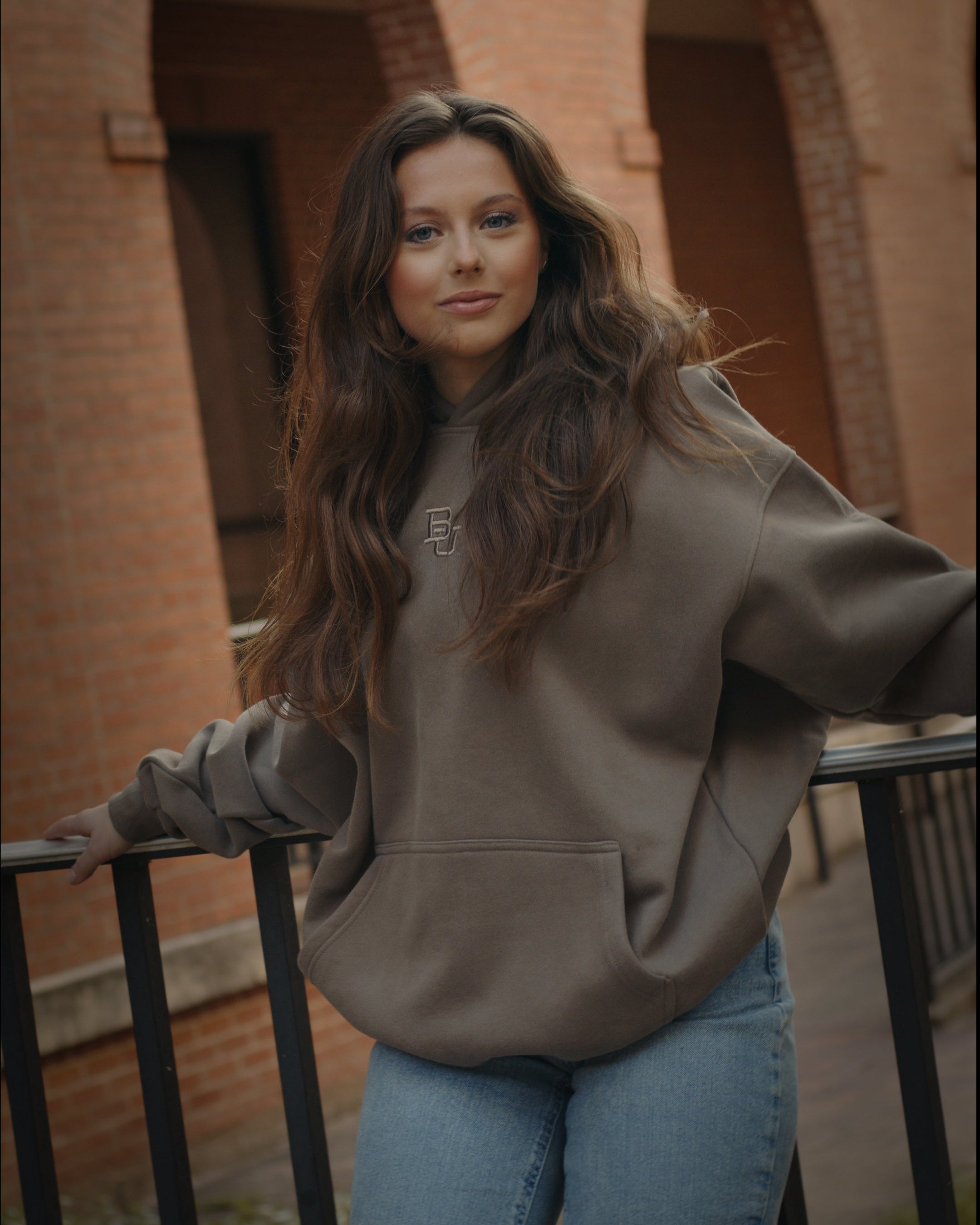 Beautiful girl wearing an olive green Baylor University mclane stadium hoodie with embroidered 'BU' in the front posing by draper and brick walls