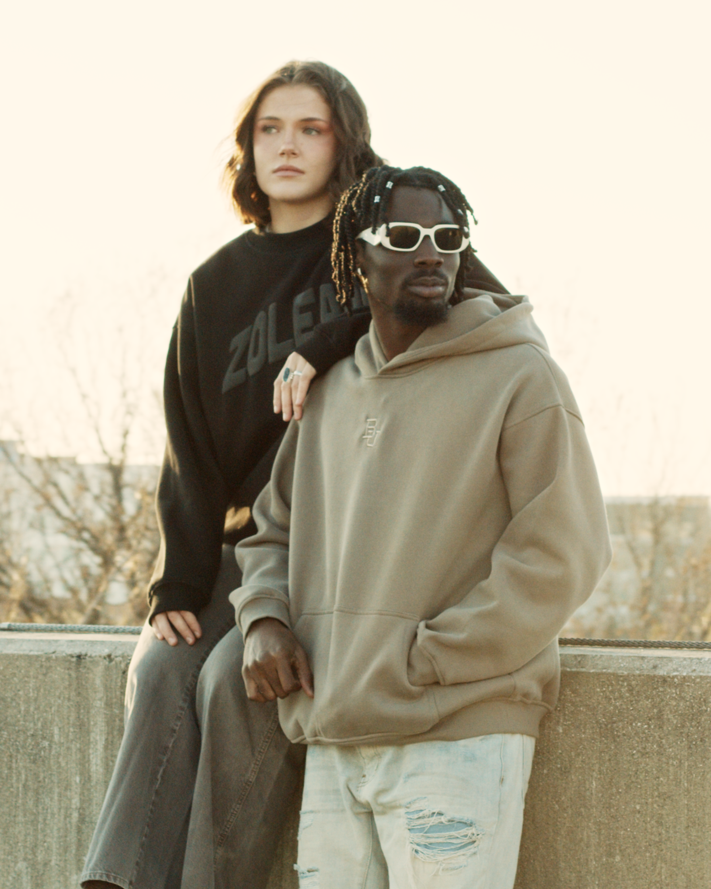 Two people standing close together on a ledge with a neutral background. Black guy with white prada sunglasses posing with white girl resting her arm on his shoulder wearing zoleado merch