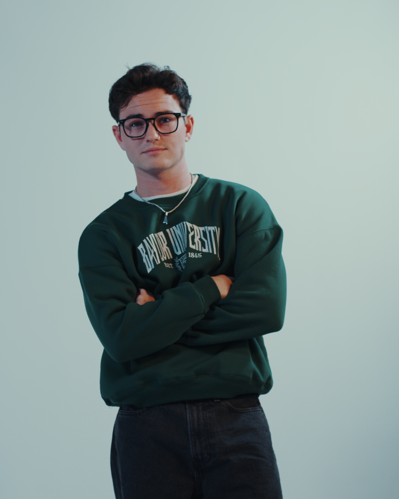 Hot male model with glasses wearing a green embroidered Baylor University sweatshirt crewneck crossing his arms on a white background