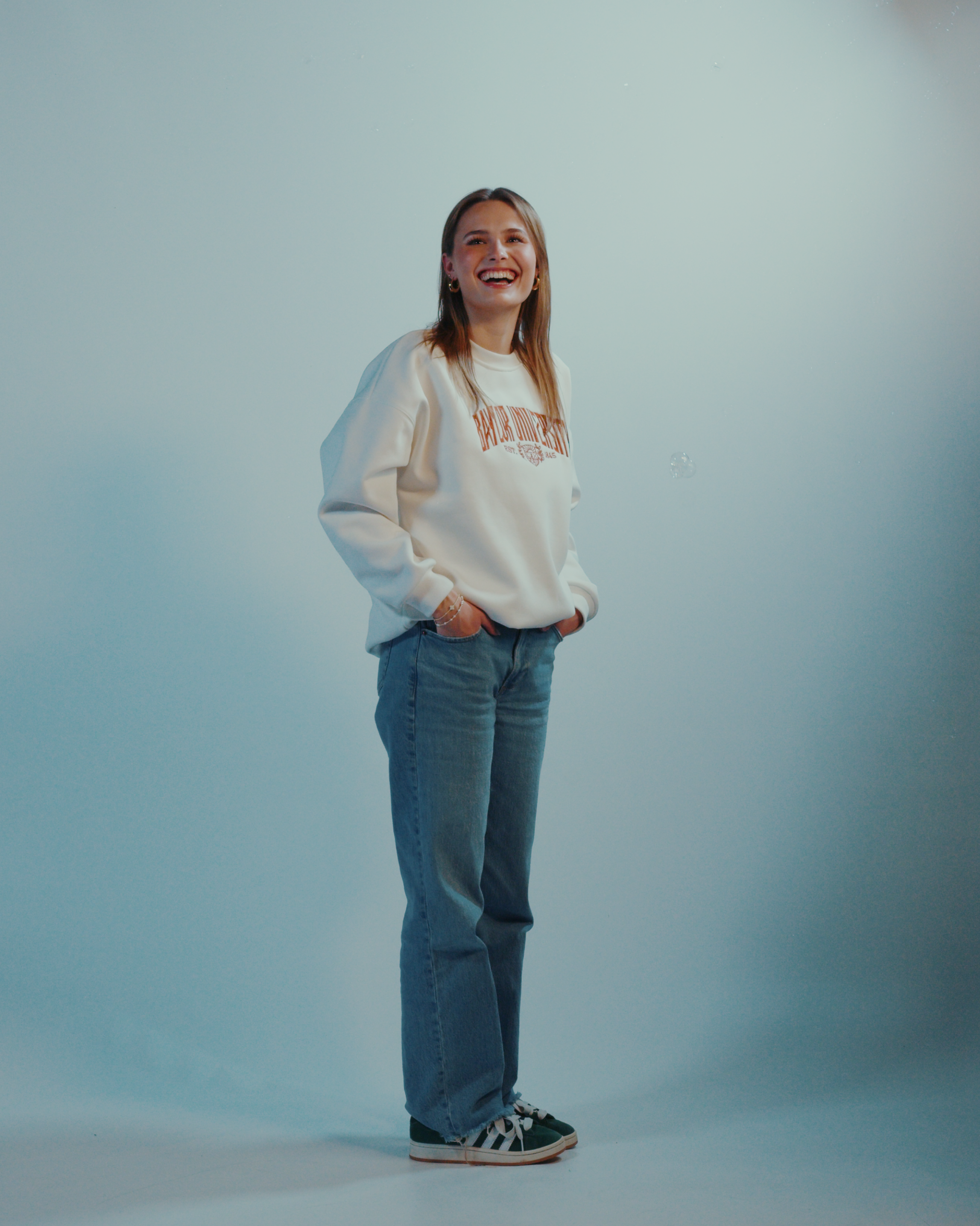 woman wearing a white Baylor University sweatshirt with text and blue jeans on a light gray background