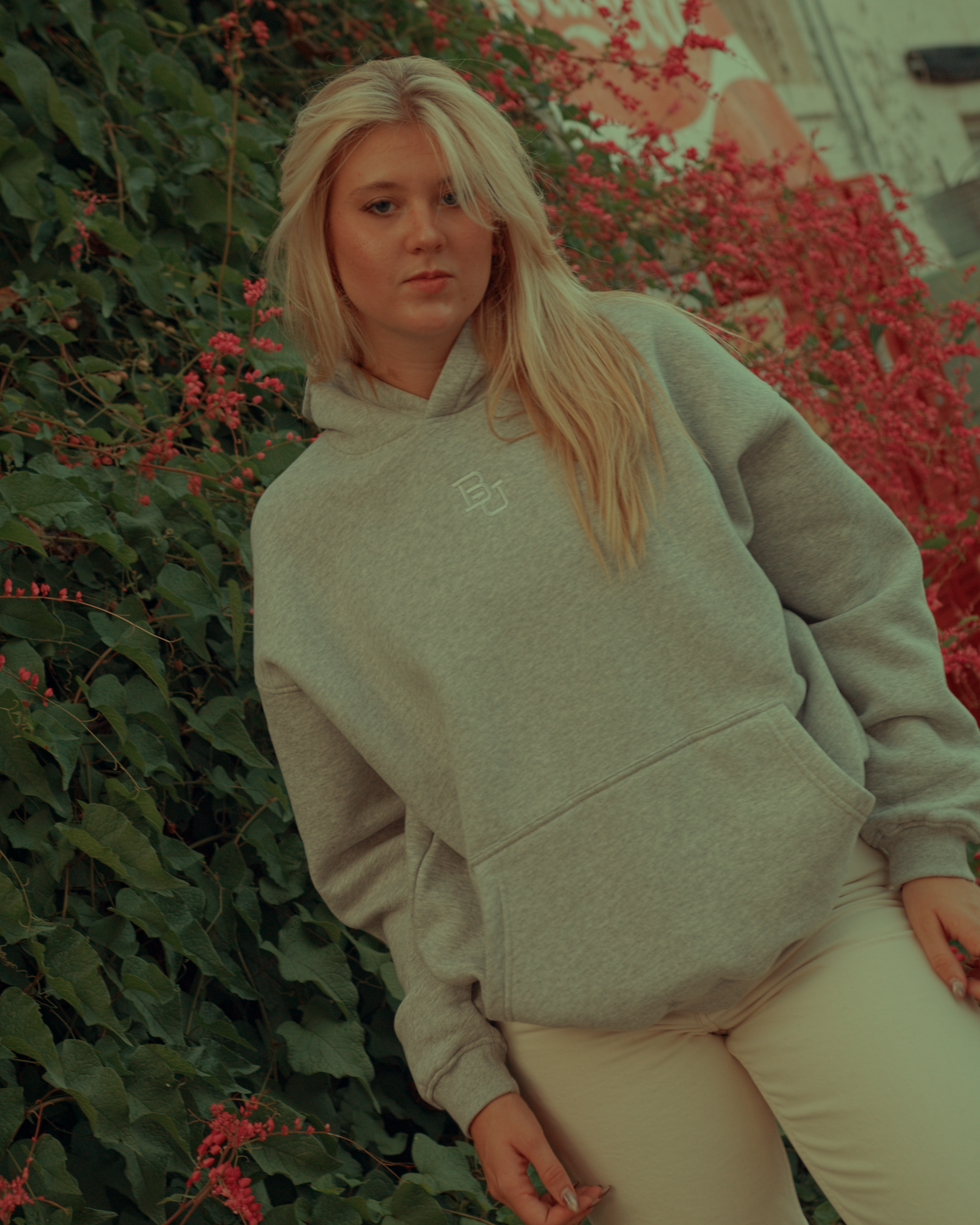 Blonde girl wearing a light gray Baylor hoodie with a logo, sitting against a green wall with red flowers.