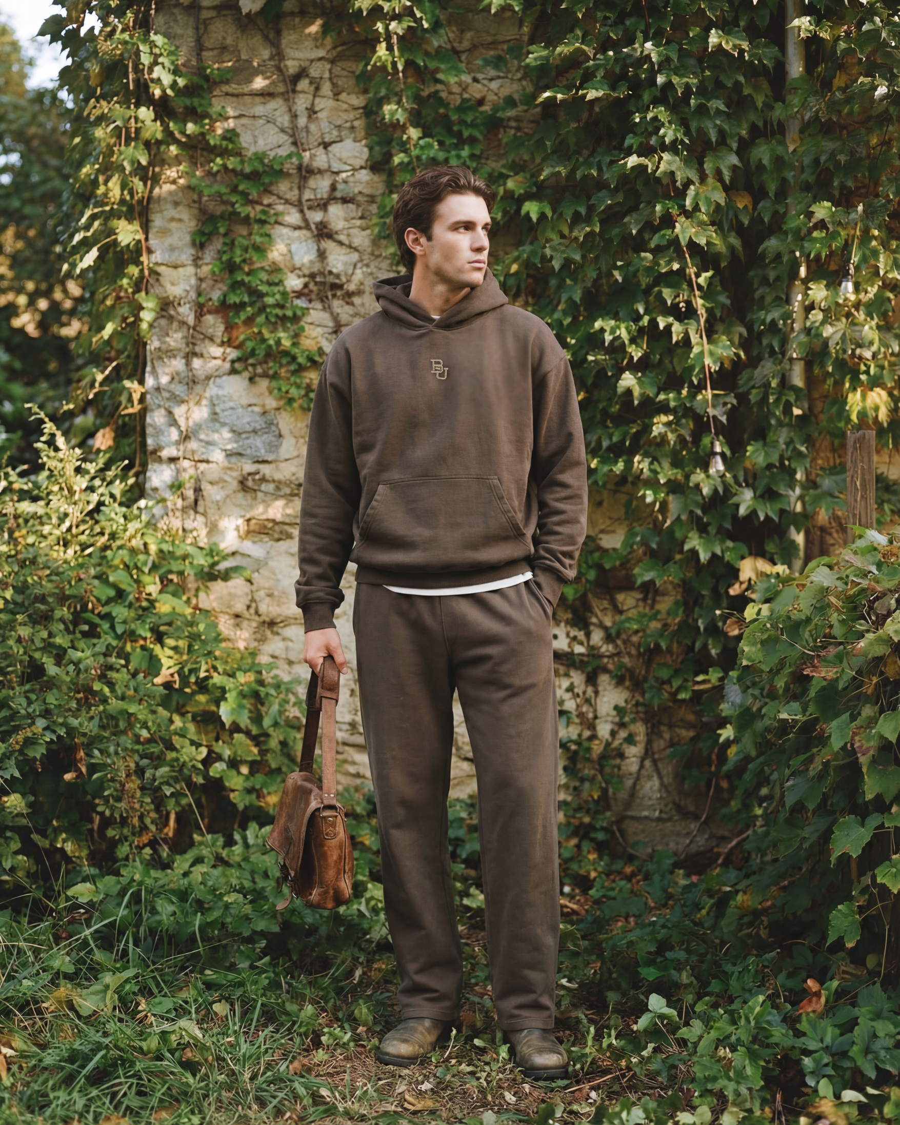 Man in brown baylor university hoodie and pants holding a leather bag in a garden setting