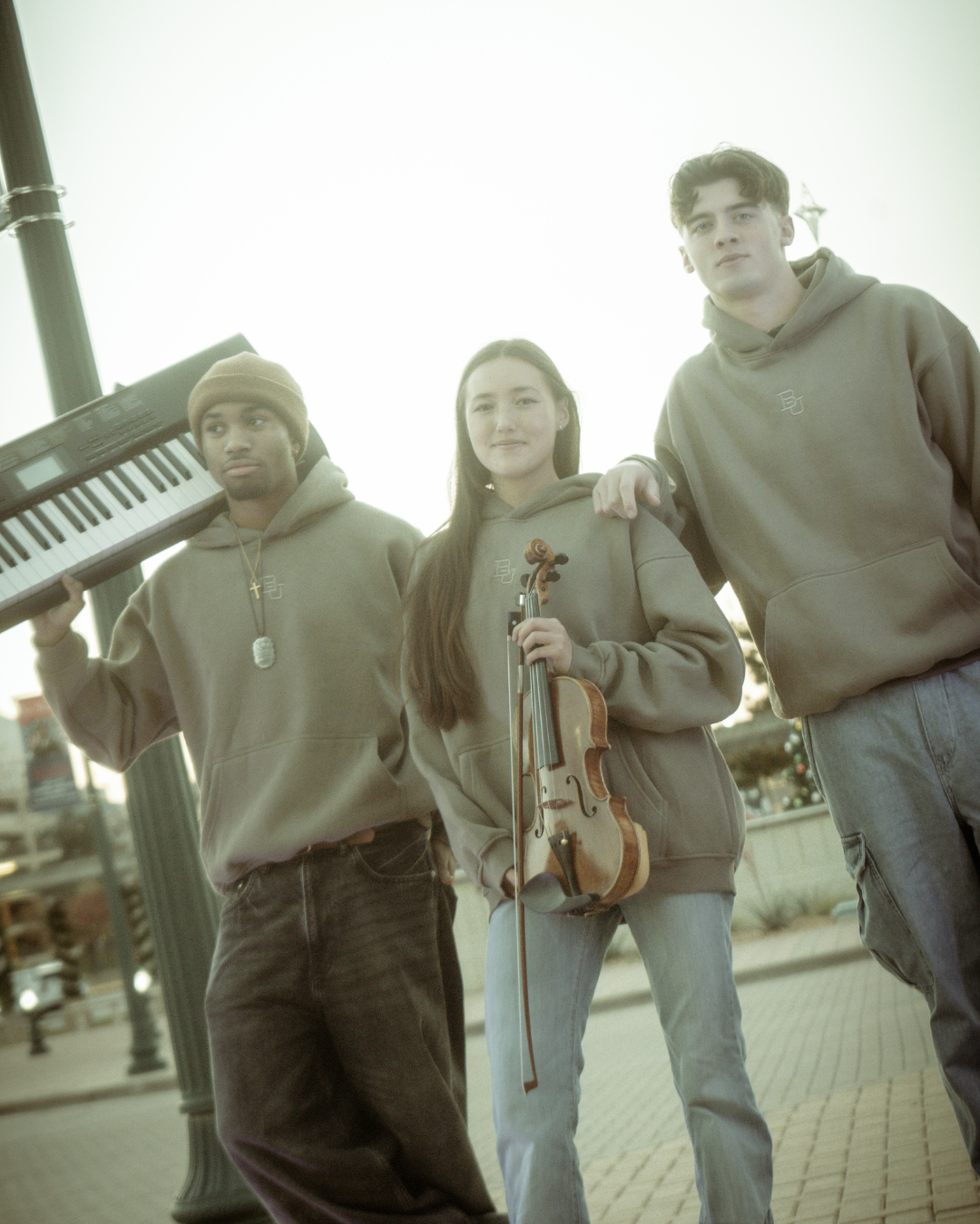 Three models wearing the baylor university mclane stadium hoodie as they hold instruments, black guy holds piano, asian girl holds a violin, white guy rest his and on the girl's shoulder