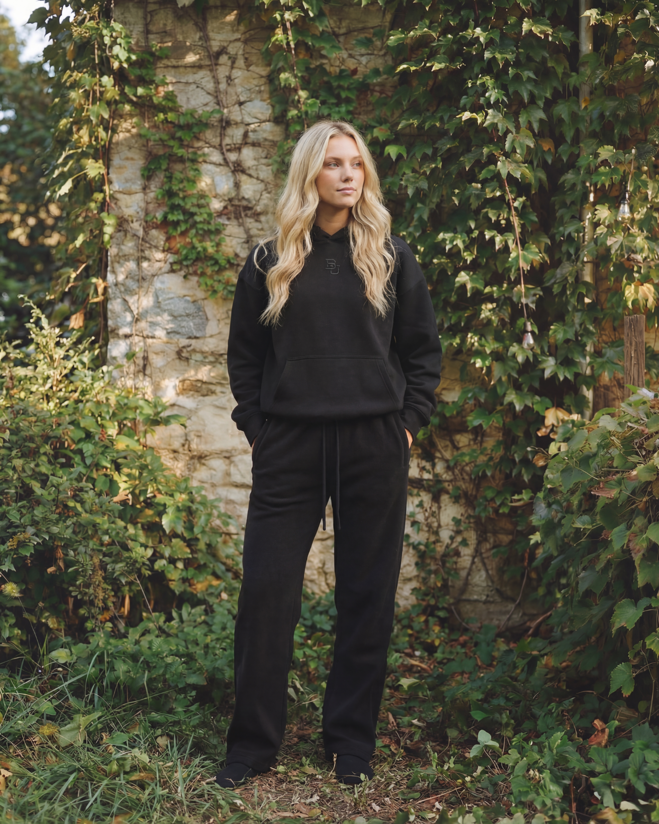 blonde woman wearing a black baylor university hoodie and sweatpants standing in a natural setting with greenery and a stone wall.
