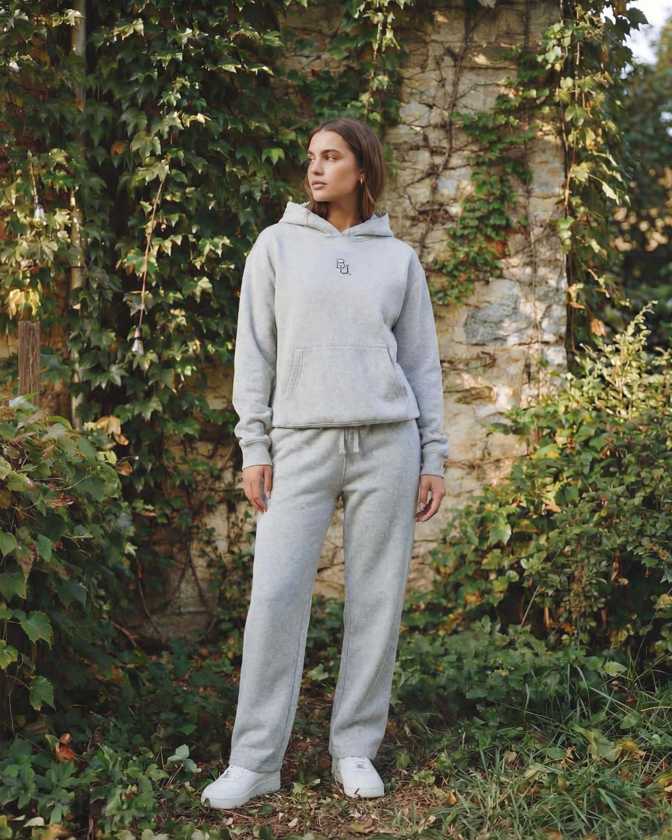 woman wearing a gray baylor hoodie and pants standing in front of a stone wall with greenery 